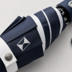 Close-up payung lipat compact premium warna navy dengan logo corporate pada panel kain, menampilkan detail tekstur bahan dan desain minimalis elegan dengan pencahayaan studio lembut.