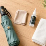 Perlengkapan perawatan payung lipat: payung compact, microfiber cloth, lubricant, dan handuk di meja kayu